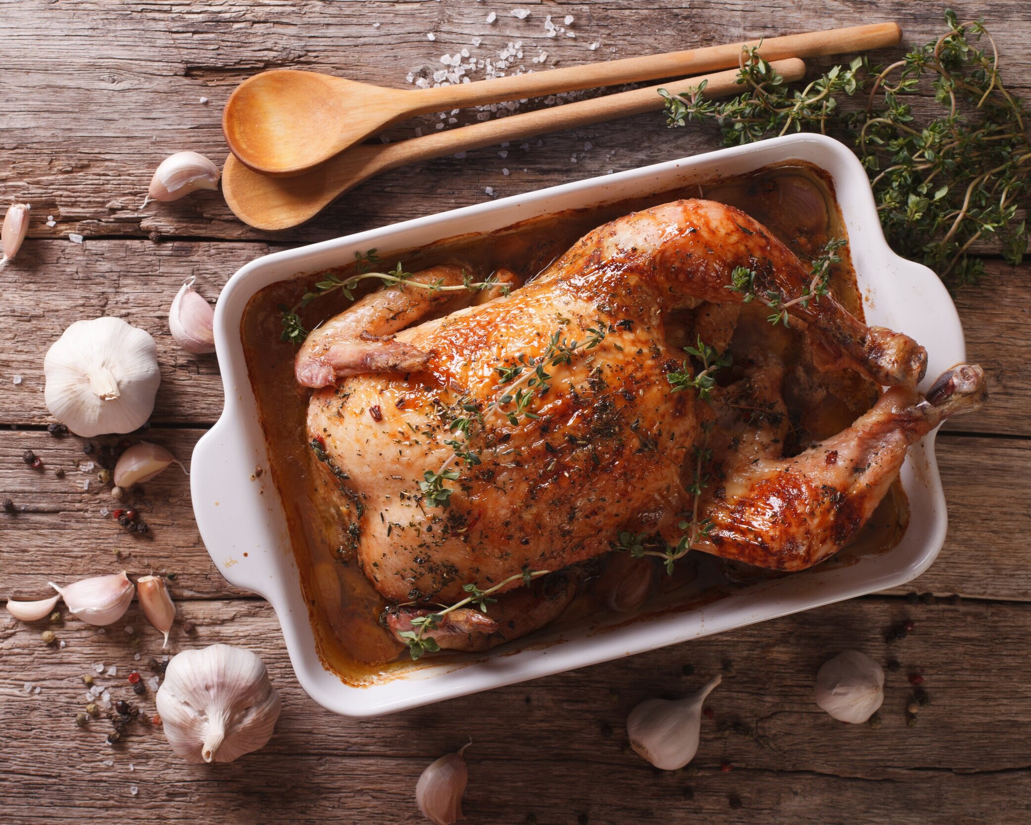 Roasted chicken garnished with fresh thyme in a white baking dish, surrounded by garlic cloves and wooden utensils on a rustic wooden surface. Perfect for a hearty meal or festive gathering.